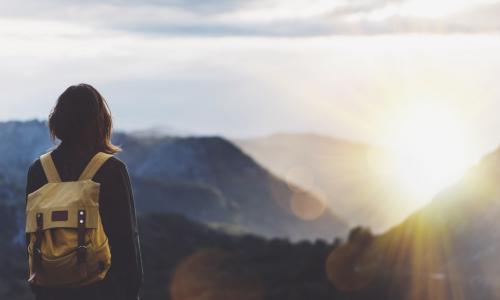 woman with backpack gazing toward sunset over mountain peaks