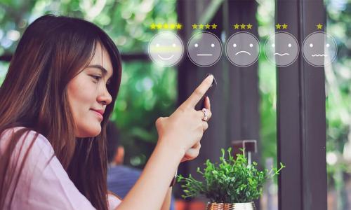 young woman with long brown hair sitting at café counter using smartphone to rate experience using scale of smiley faces ranging from happy to unhappy
