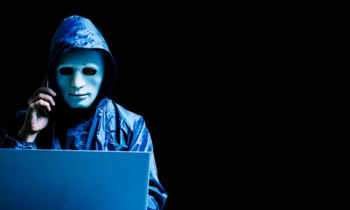 scammer in mask and hoodie with phone and laptop