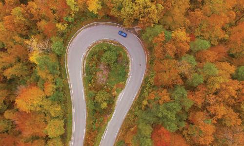 blue car driving along sharp hairpin turn in the road in the middle of a forest with autumn leaves