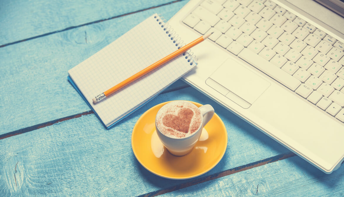 coffee with a heart in the milk on a teal table next to a notebook and a laptop