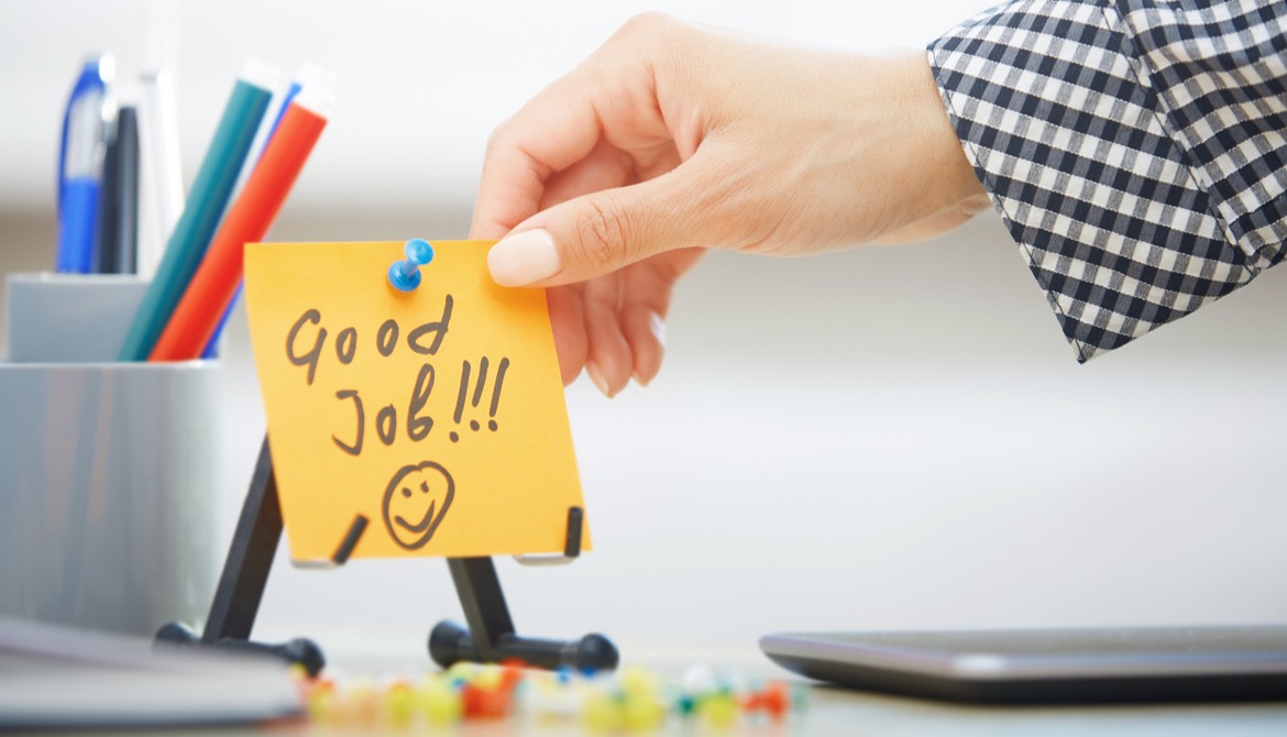 manicured hand reaching out to touch yellow good job sticky note on work desk