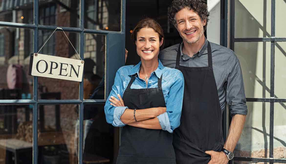 husband and wife small business owners stand outside their shop with open sign in window