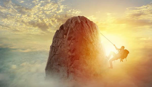 A mountain climber about to reach the top of a mountain peak in front of a cloudy sunset sky 