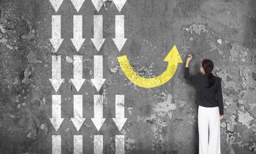 woman in front of black board with lots of white arrows pointing straight down and one yellow arrow curving around to point up, representing making a different decision