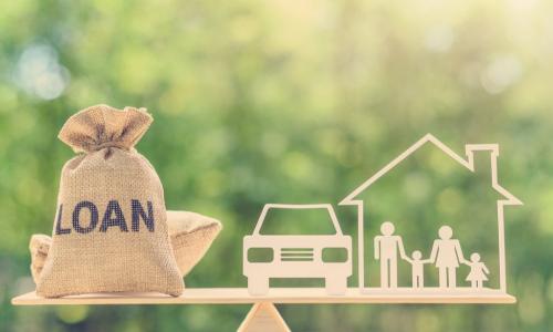 burlap bag with loan written on it balancing with a car and house on a wooden balance