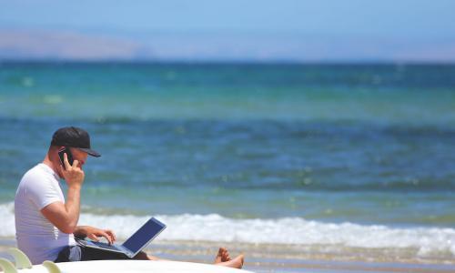 Many works or learns on a laptop at the beach next to a surf board