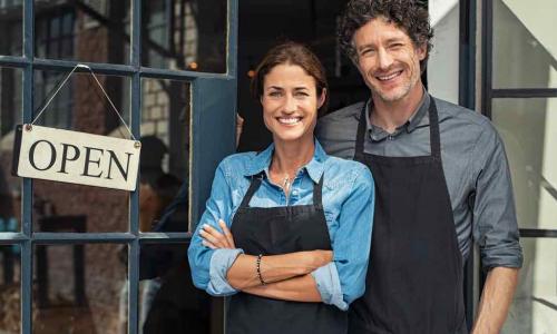 husband and wife small business owners stand outside their shop with open sign in window