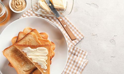 stack of buttered toast on plate next to butter knife and bowls of butter and peanut butter